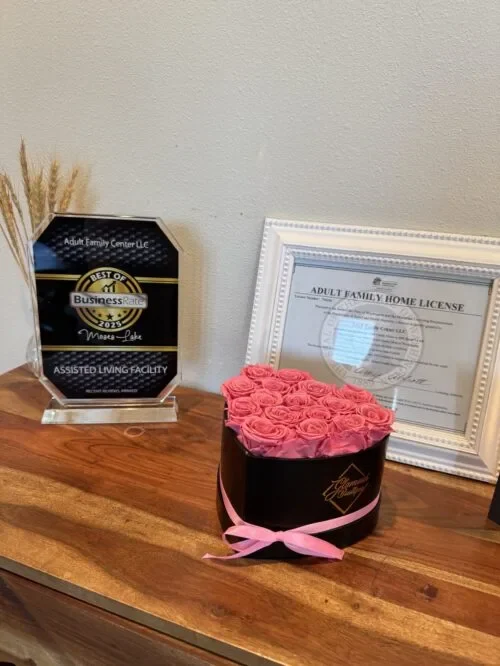 A black round box with pink preserved roses and a pink ribbon sits on a wooden table beside a black plaque and framed certificate.